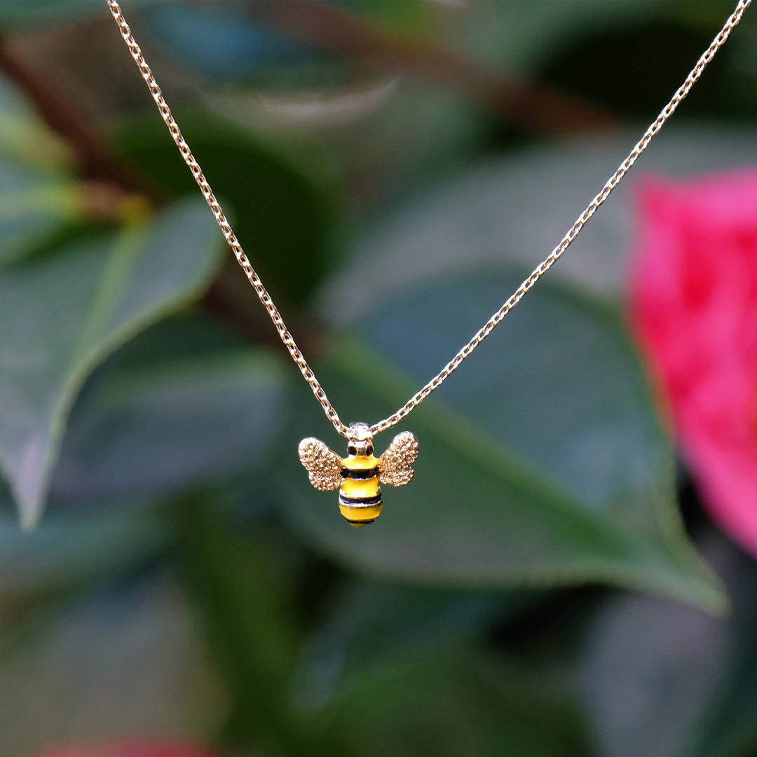 Gold bee-shaped pendant necklace against a blurred green and pink floral background