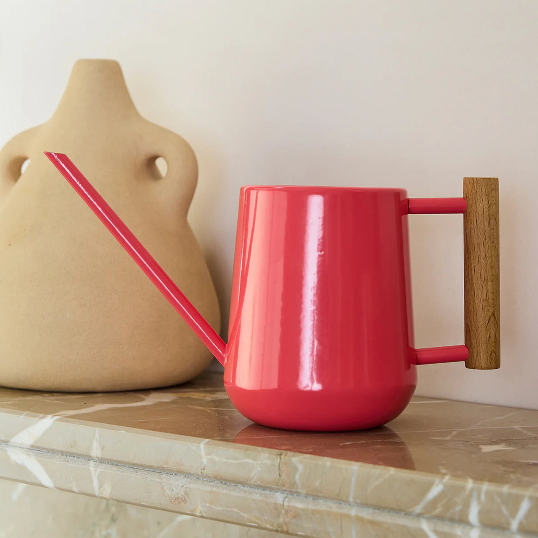 Coral watering can with a wooden handle on a marble surface next to a beige vase.