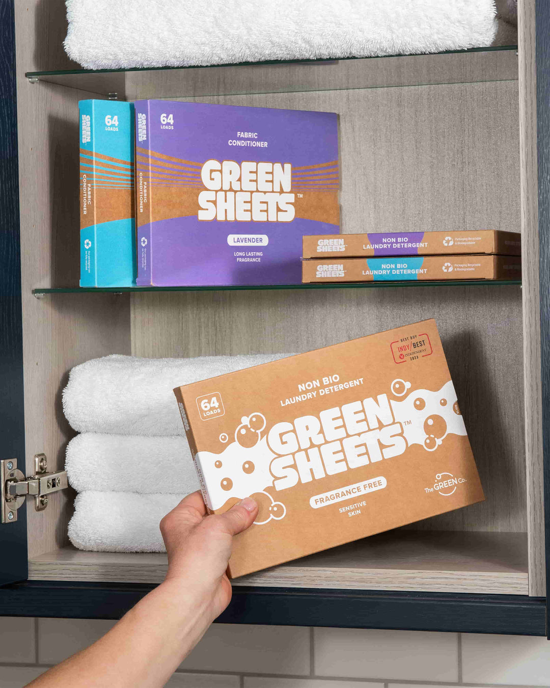 Hand holding a box of Green Sheets laundry detergent in front of shelves with folded towels and more boxes.