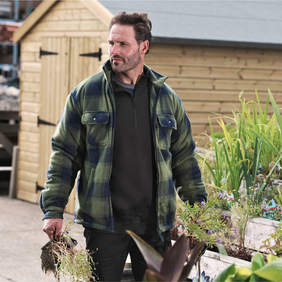 Man in a plaid jacket walking outdoors with plants, wooden shed in the background