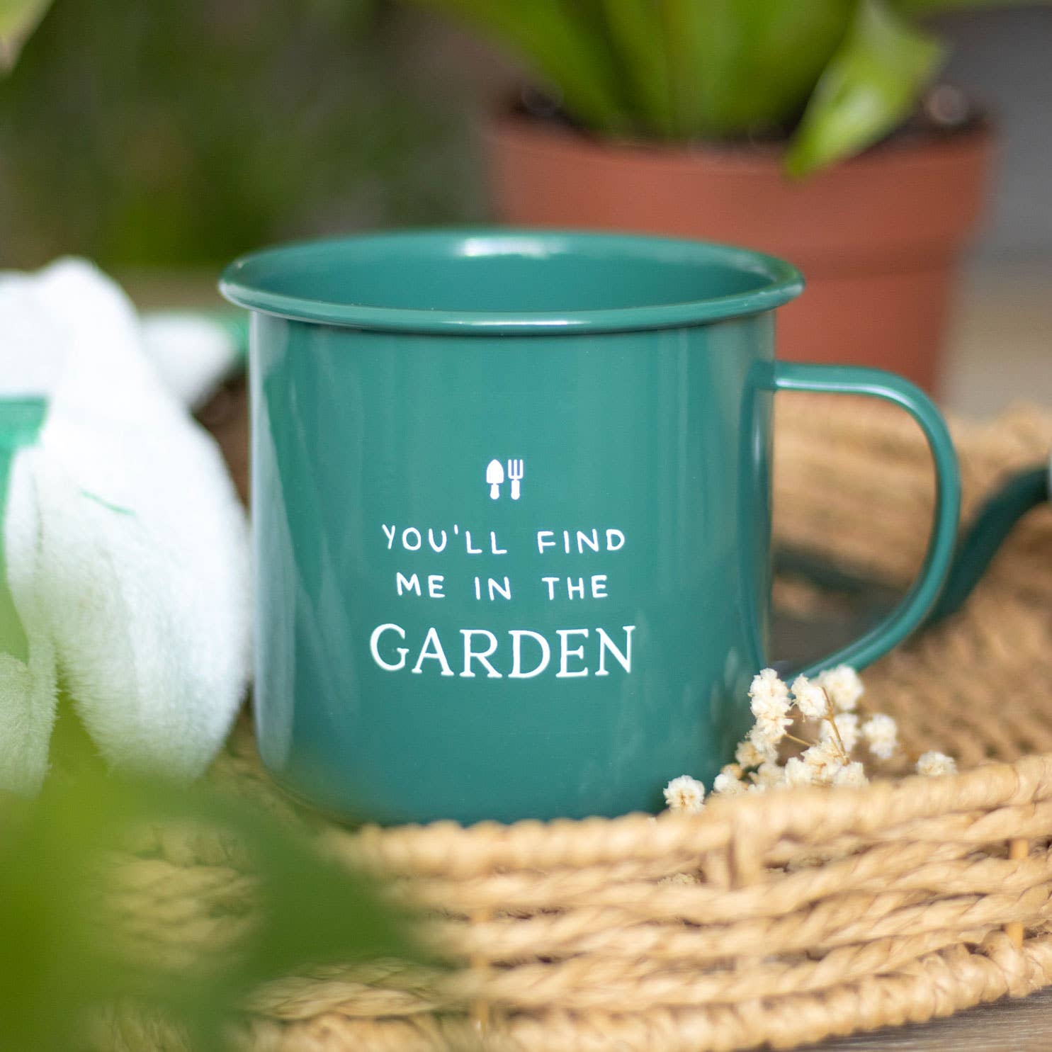 Green mug with 'You'll find me in the garden' text on a woven mat with plants in the background
