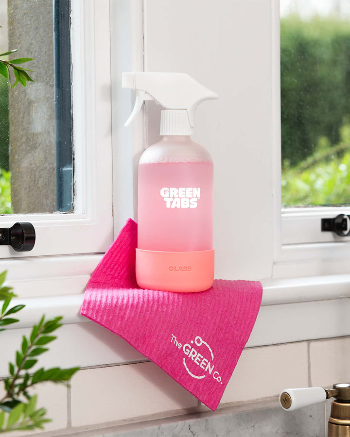 Pink spray bottle with white nozzle on a pink towel on a windowsill with greenery in the background