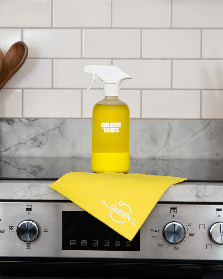 Yellow spray bottle and cloth on a stove with 'Green Gains' branding against a tiled wall background.