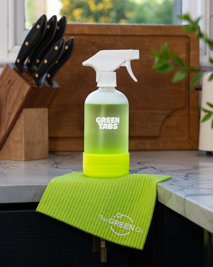Green Tabs cleaning spray bottle and towel on a kitchen counter with window and plant in background