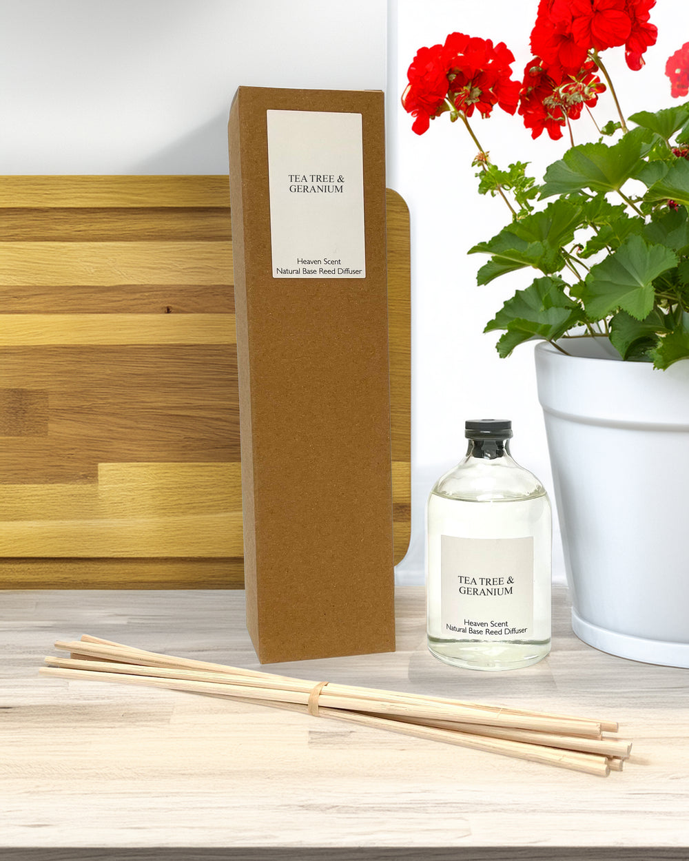 A glass bottle and reeds as part of a reed diffuser kit with box on a counter top with red flowered geranium