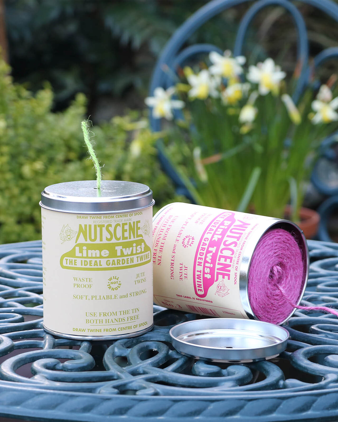 Two Nutscene twine tins, one lime twist and one pink, on a garden table with flowers in the background.