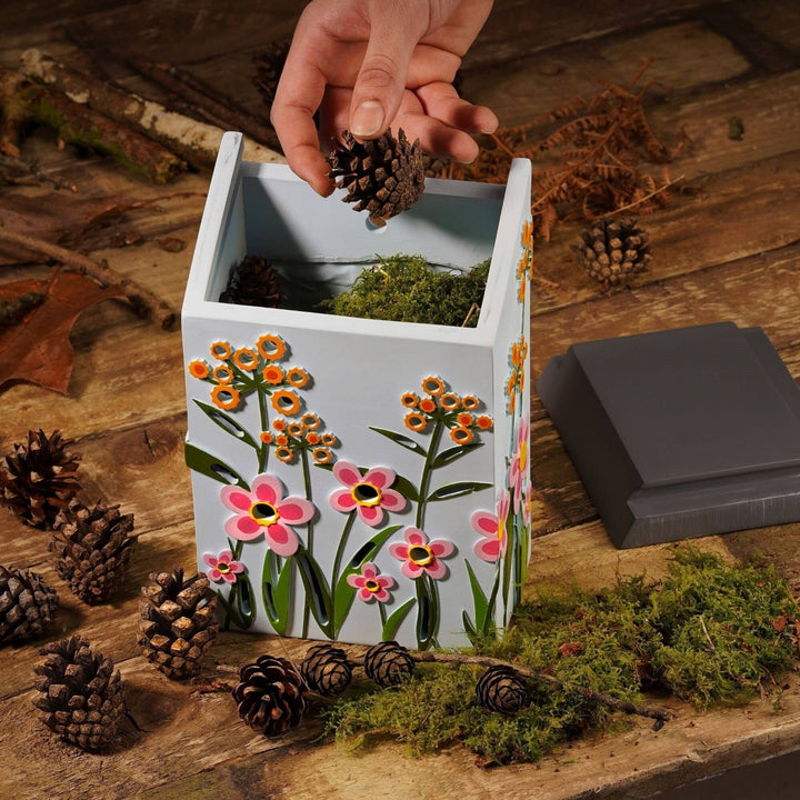 Decorative insect house with floral design on a wooden surface with pinecones and moss.