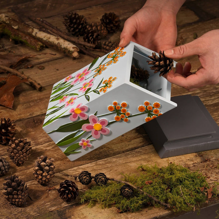 Hand placing a pine cone into a small insect box with floral design on a wooden surface.
