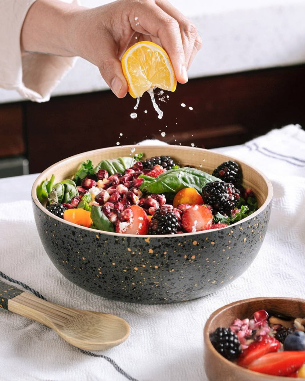 Person squeezing lemon over a salad in a speckled bowl on a white tablecloth.