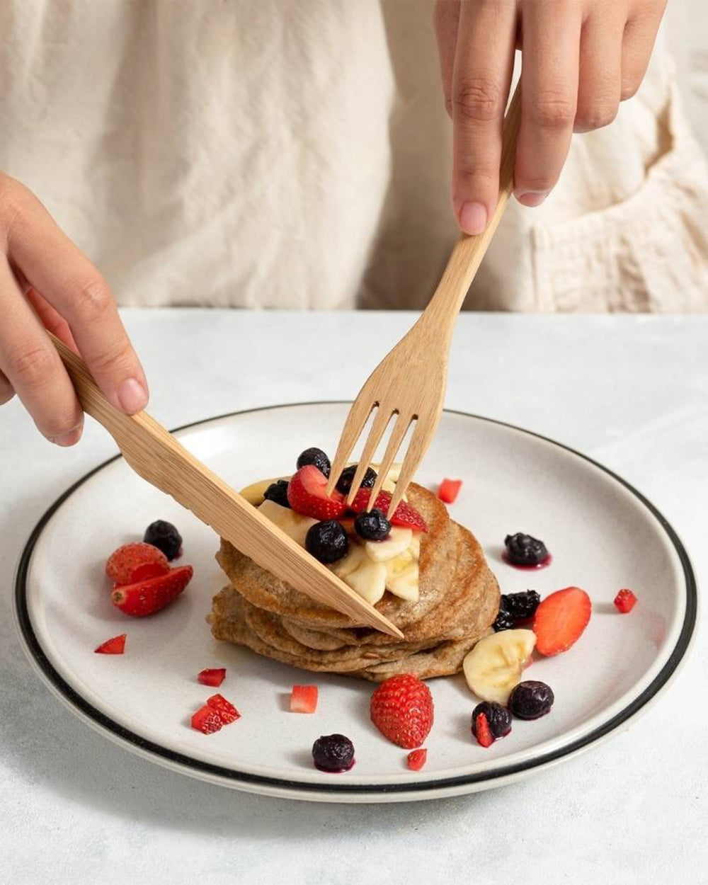 Stack of pancakes with fruit on a plate, being cut into with wooden utensils.