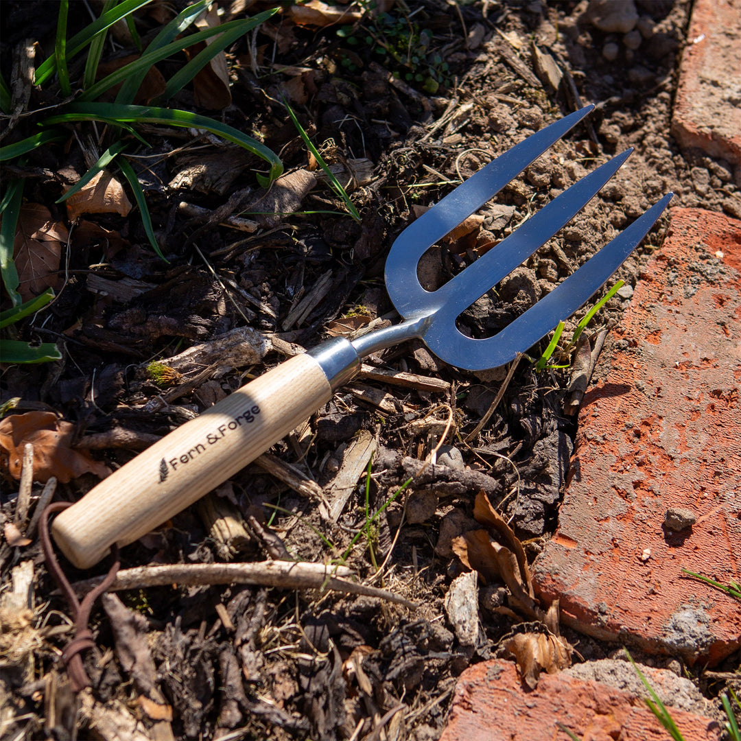 Nutscene Fern and Forge hand fork with stainless steel tines and ash handle resting on soil beside a brick garden edge