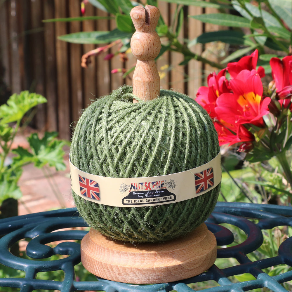 A green ball of yarn wound on a wooden spool with a small wooden top, sitting on a teal metal garden table outdoors. A paper label around the yarn reads 'Nutscene' and features Union Jack flags.