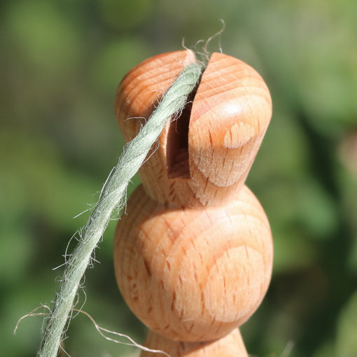 A close-up of a light natural wood spoindle copntaining a twine cutting notch, holding a green garden twine, with a blurred outdoor garden background featuring red flowers.