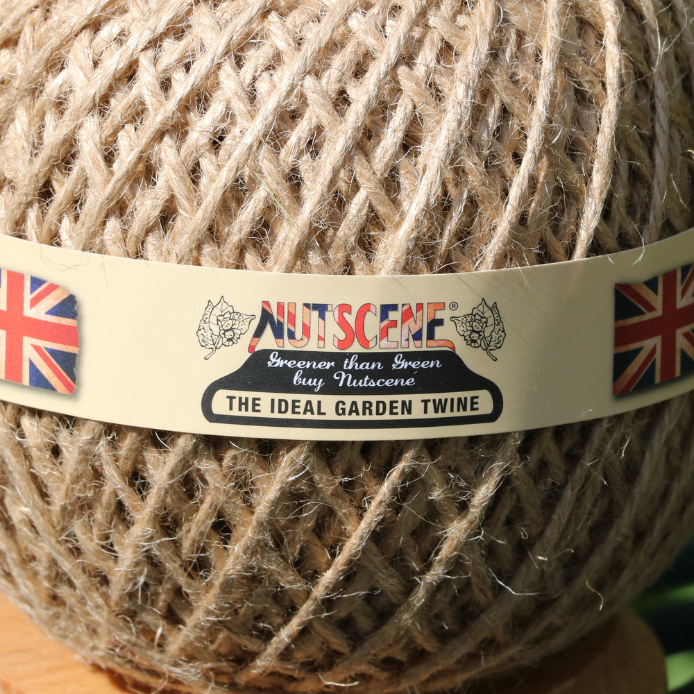 A close-up of a ball of natural jute garden twine wrapped with a cream paper band labeled 'NUTSCENE The Ideal Garden Twine' with Union Jack flags on either side, set against a blurred green outdoor background.
