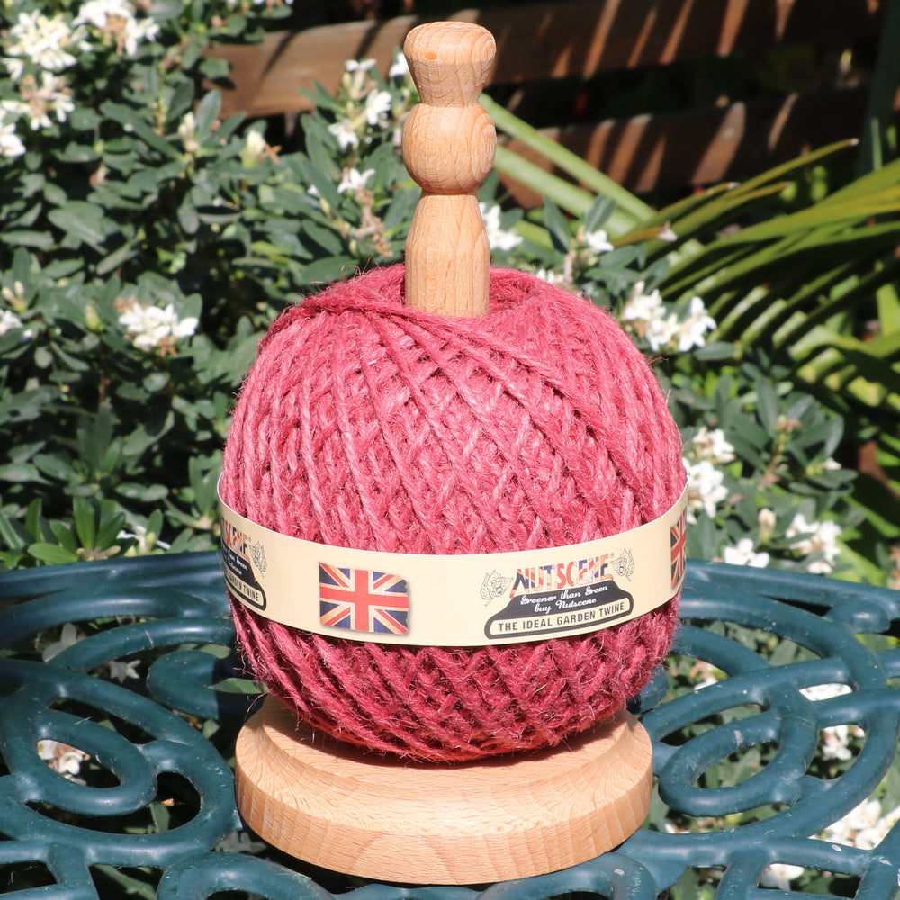 A wooden garden twine dispenser with a ball of red twine wound around it, displayed on a dark green metal garden table with plants and white flowers in the background.
