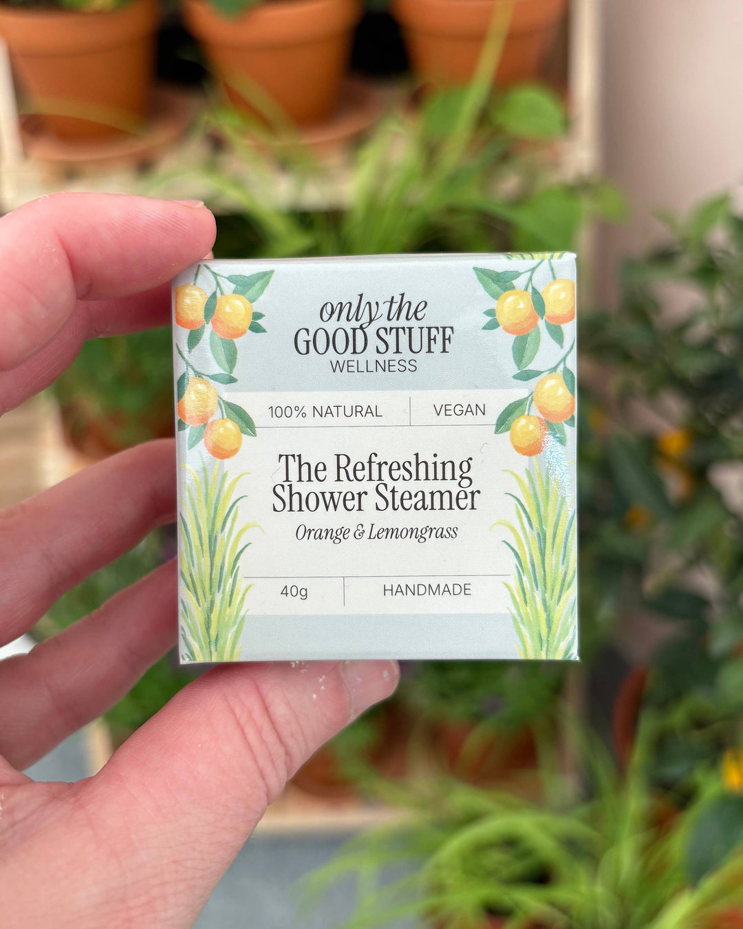 Hand holding a box of 'Only the Good Stuff' shower steamer with plants in the background