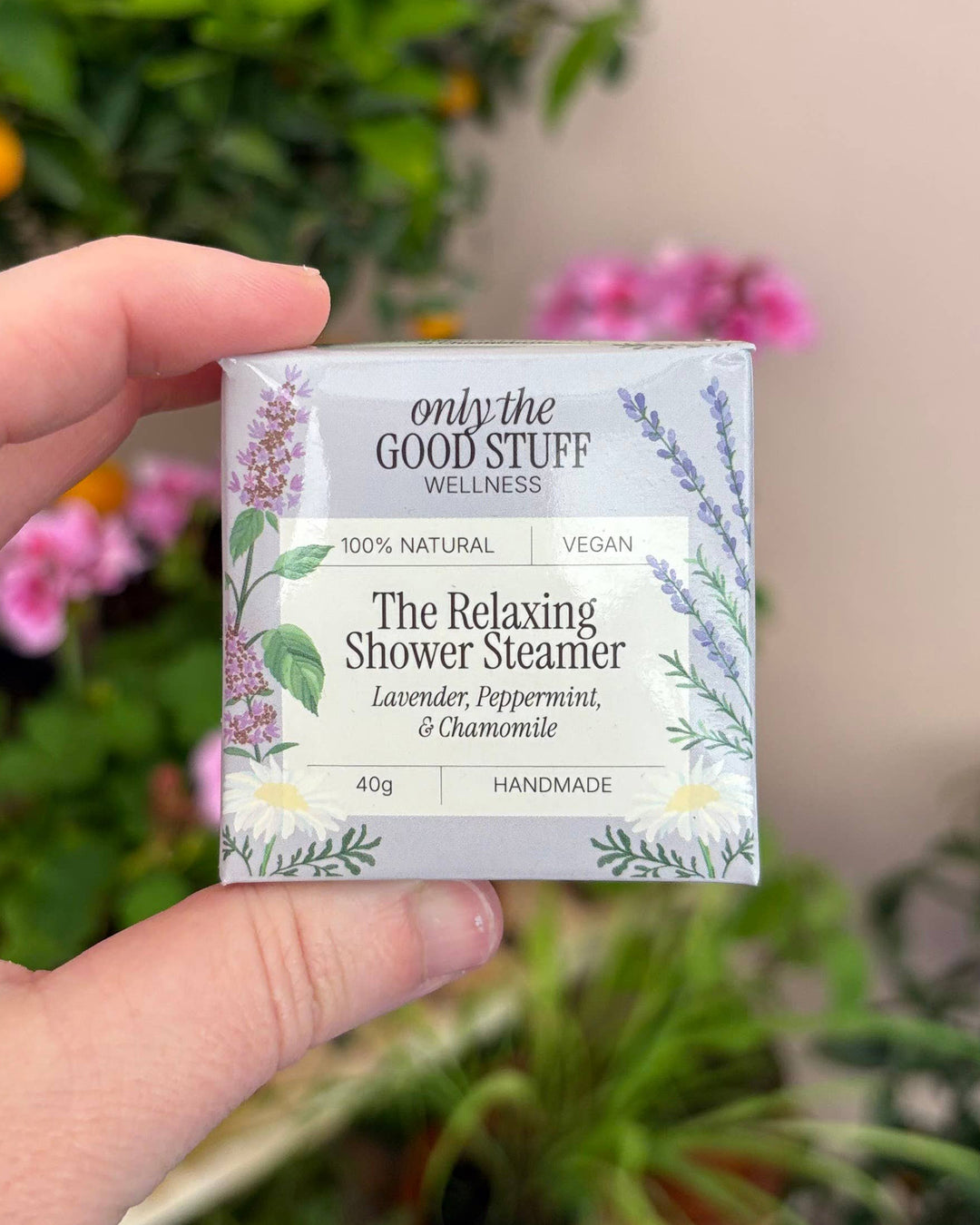 Hand holding a box of 'only the Good Stuff' Relaxing Shower Steamers with a garden background.