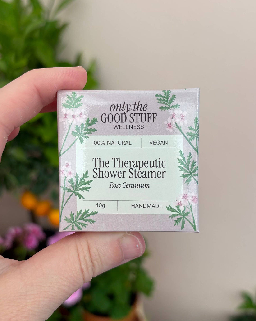 Hand holding a box of 'only the Good Stuff Wellness' Therapeutic Shower Steamer with plants in the background