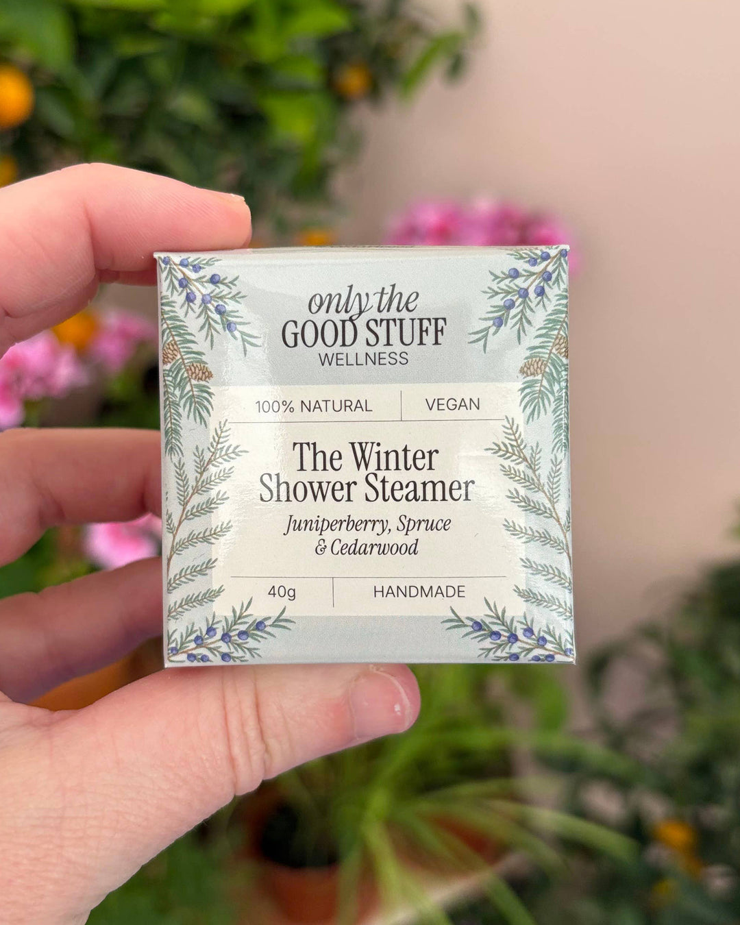 Hand holding a 'only the Good Stuff' winter shower steamer packaging with a blurred floral background.