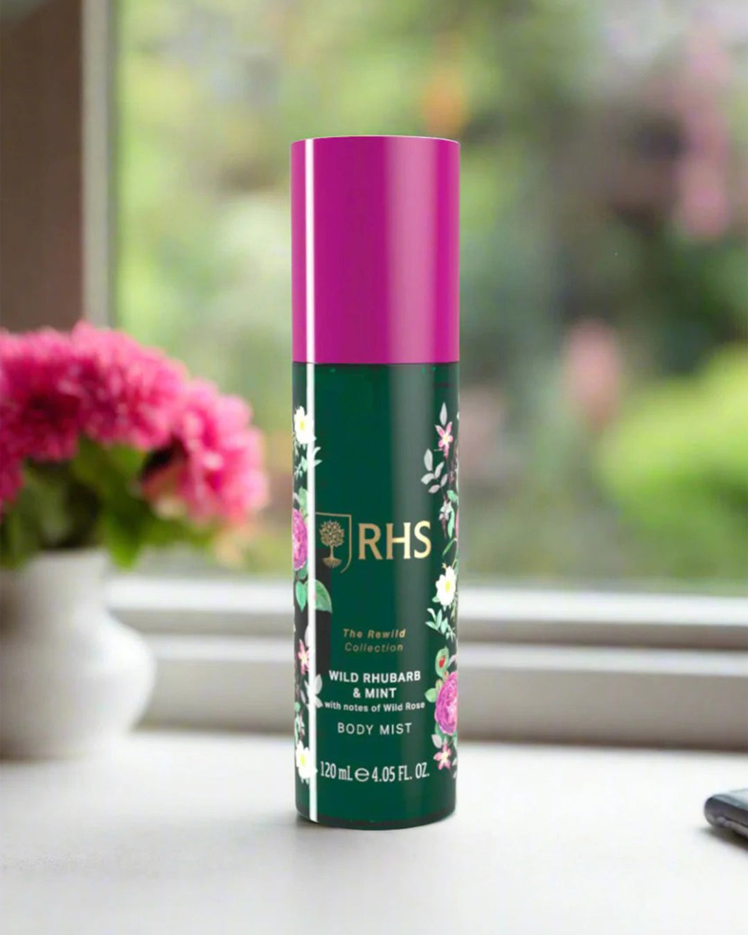 RHS body mist bottle with pink cap on a surface near a window with flowers.