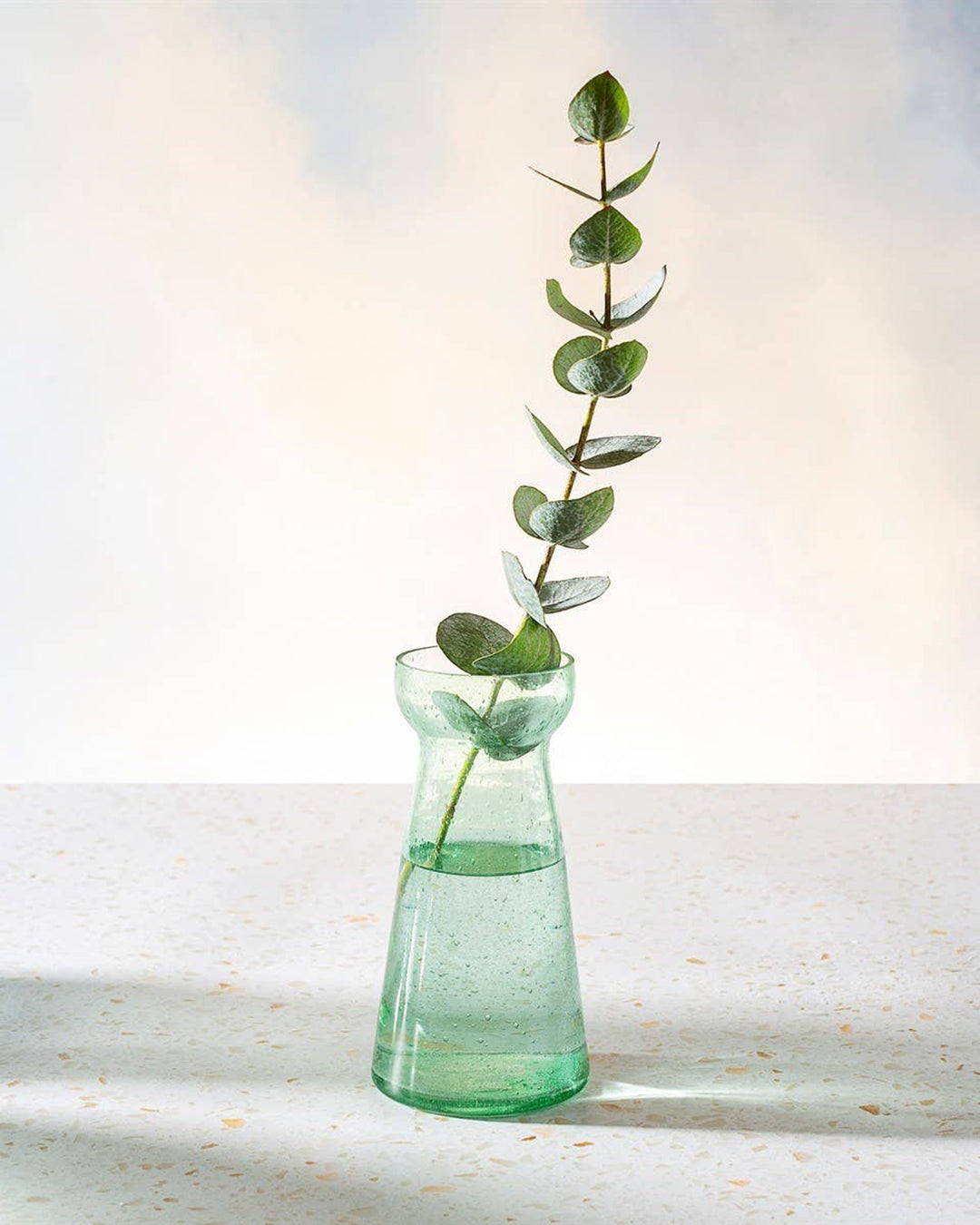 Green glass vase with a single eucalyptus branch on a light background