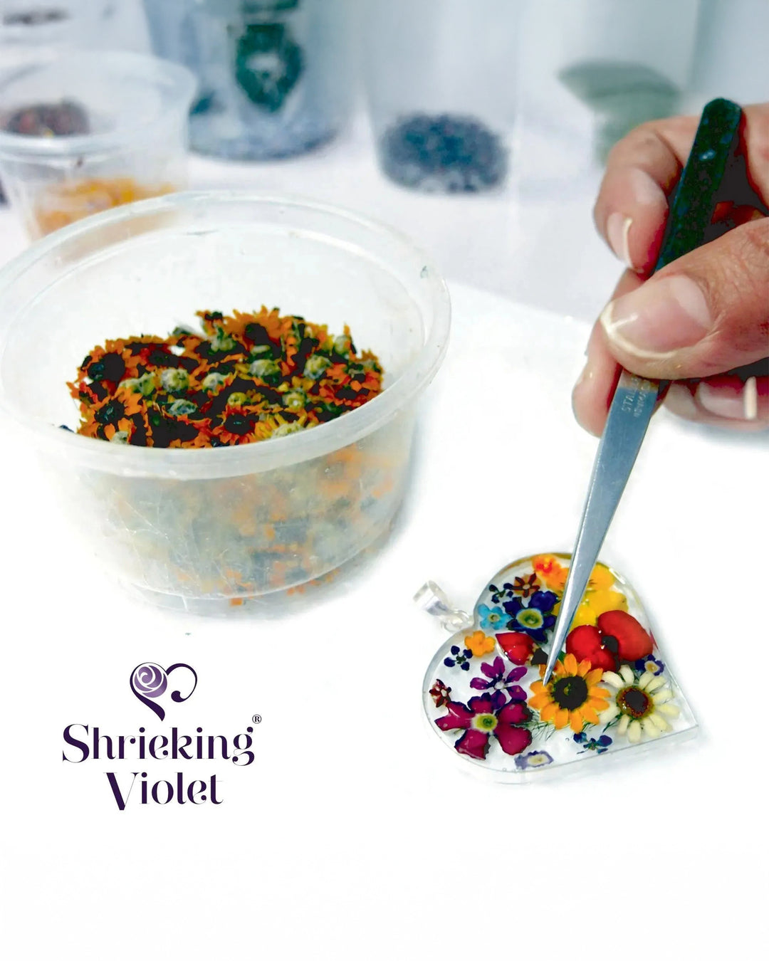 Colorful flower mix in a clear container with a hand holding a spoon, next to a heart-shaped mold with flowers, on a white surface. 'Shrieking Violet' brand visible.