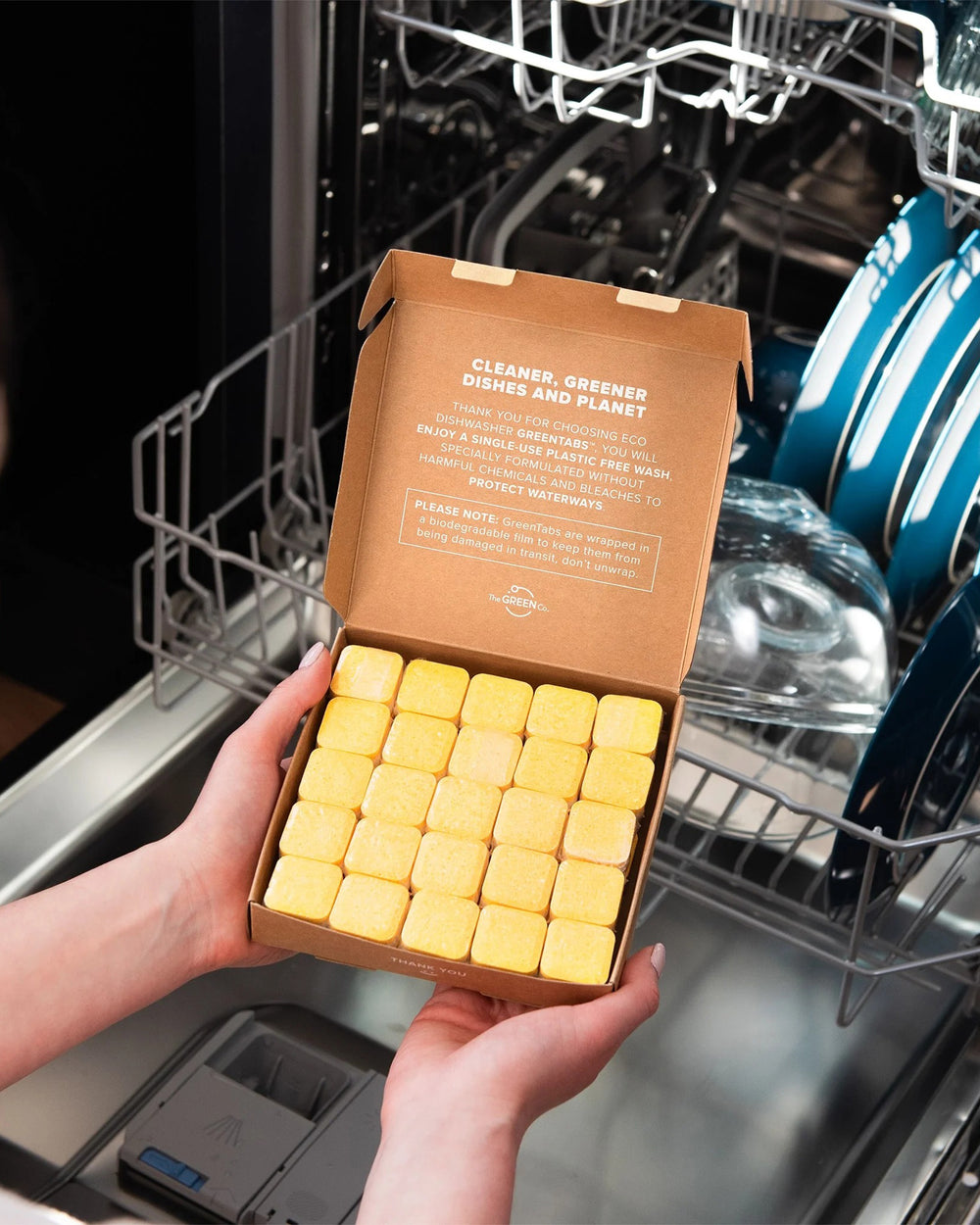 Box of yellow dishwasher pods held in front of an open dishwasher.