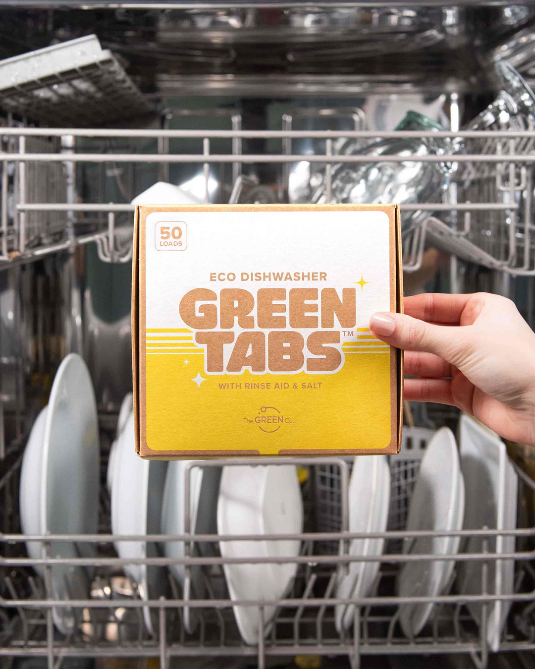 Box of Eco Dishwasher Green Tabs held in front of a dishwasher with clean dishes inside.