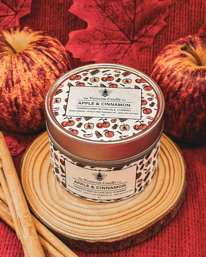 Candle jar labeled 'Apple & Cinnamon' with autumn-themed design on a red fabric background.