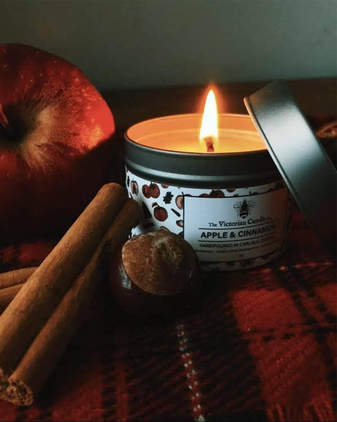 Candle with 'Apple & Cinnamon' label surrounded by cinnamon sticks and an apple on a checkered cloth.