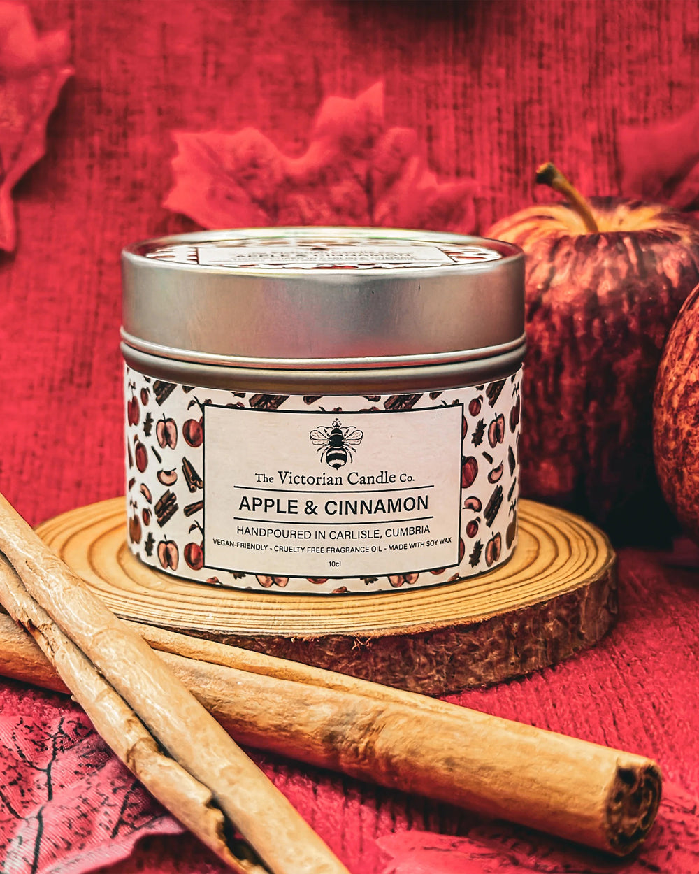 Candle tin labeled 'Apple & Cinnamon' by The Victorian Candle Co. on a red fabric background with cinnamon sticks.