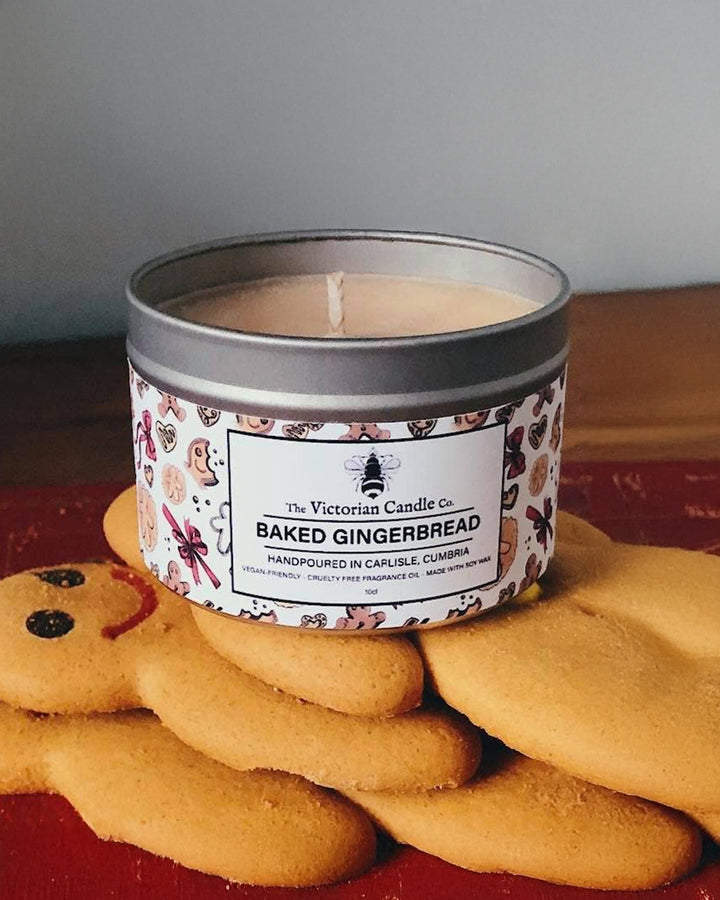 Candle in a decorative tin labeled 'Baked Gingerbread' with gingerbread cookies on a red surface.