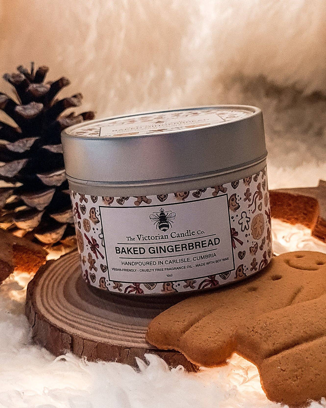 Candle in a decorative tin labeled 'Baked Gingerbread' from The Victorian Candle Co., surrounded by gingerbread cookies and a pine cone.