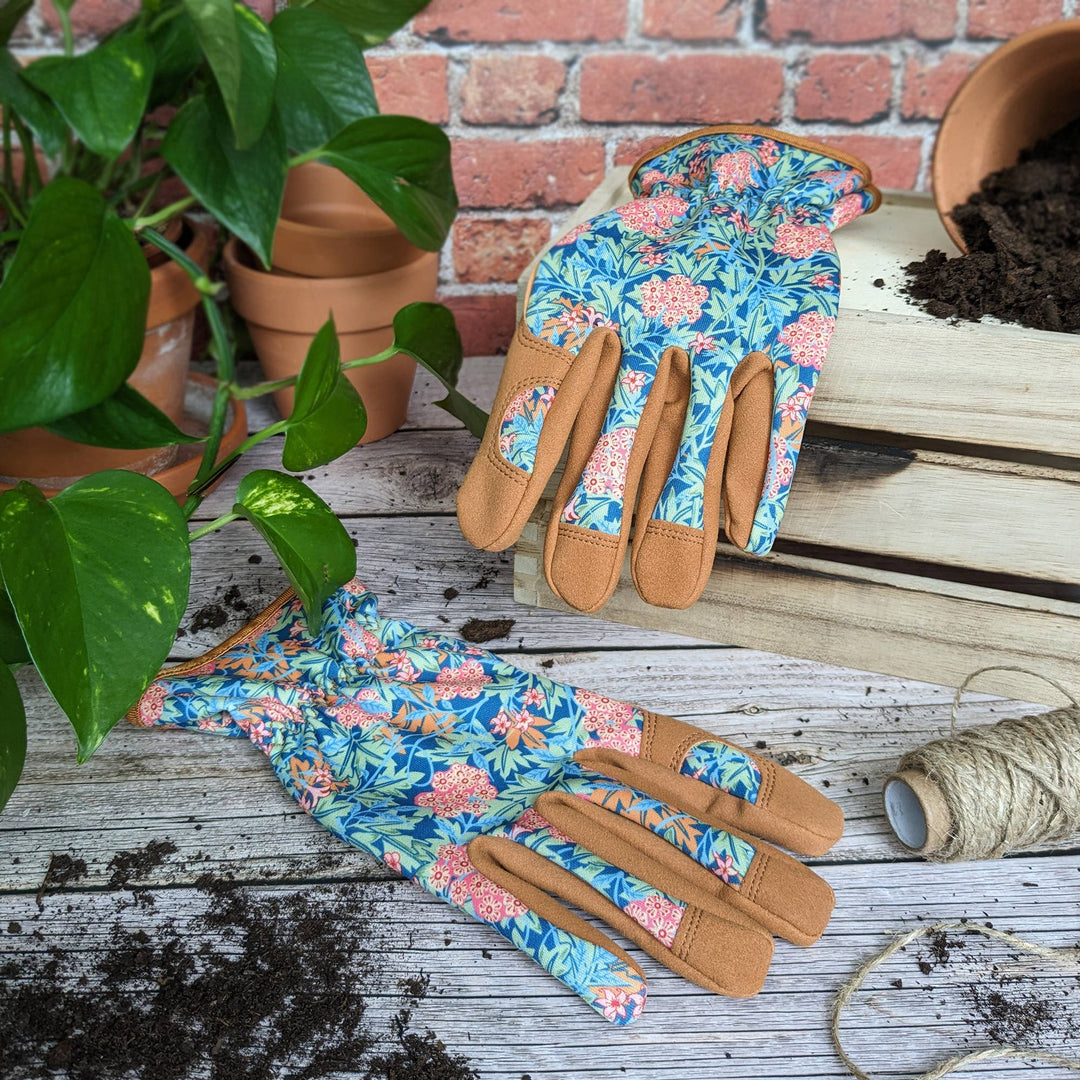 Floral gardening gloves on a wooden surface with plants and pots in the background.