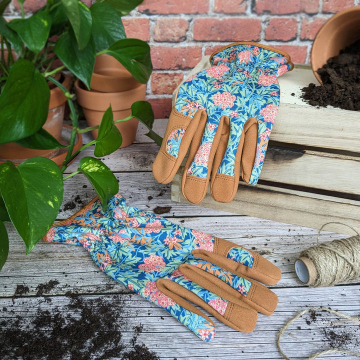 Floral gardening gloves on a wooden surface with plants and pots in the background.