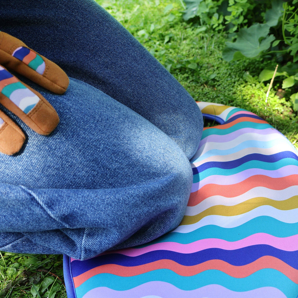 Person wearing blue jeans and colorful gardening gloves sitting on a striped mat outdoors.