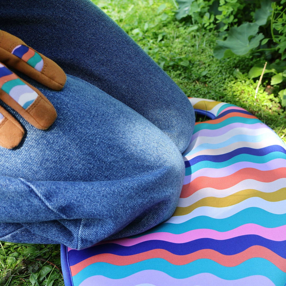 Person wearing blue jeans and colorful gardening gloves sitting on a striped mat outdoors.