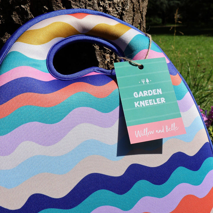 Colorful garden kneeler with a tag labeled 'Garden Kneeler' by Willow and Belle, leaning against a tree.